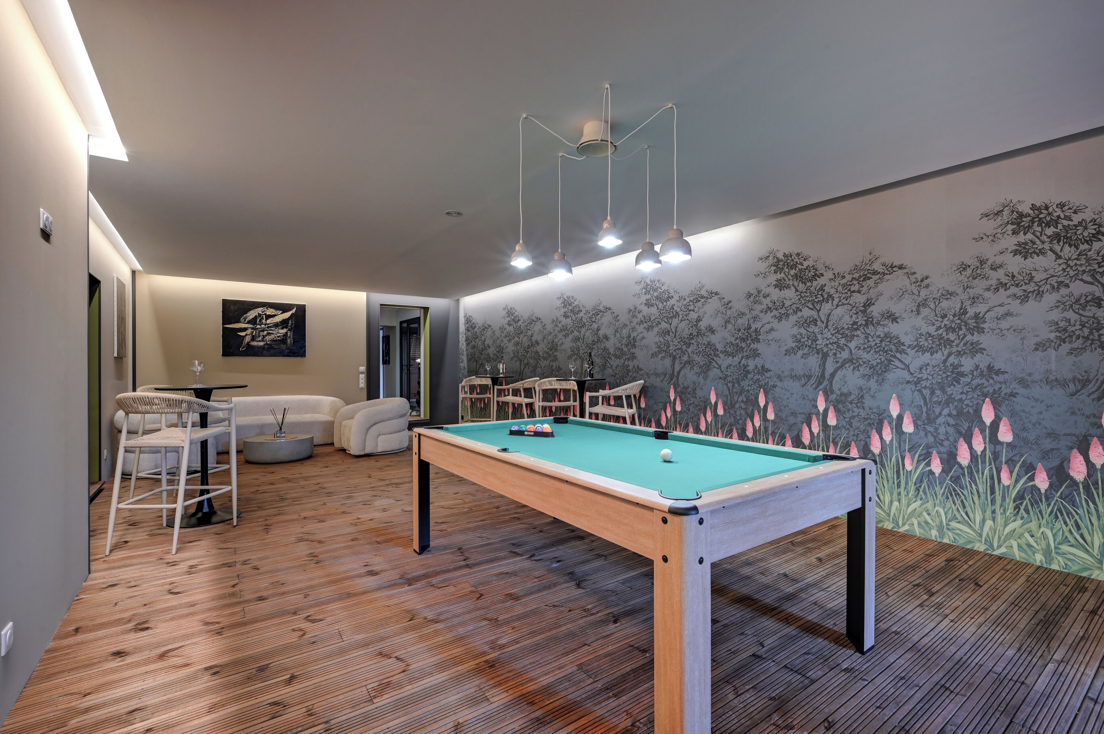 Una mesa de billar en una habitación con suelo de madera, un mural de árboles y flores en la pared y una lámpara en el techo.