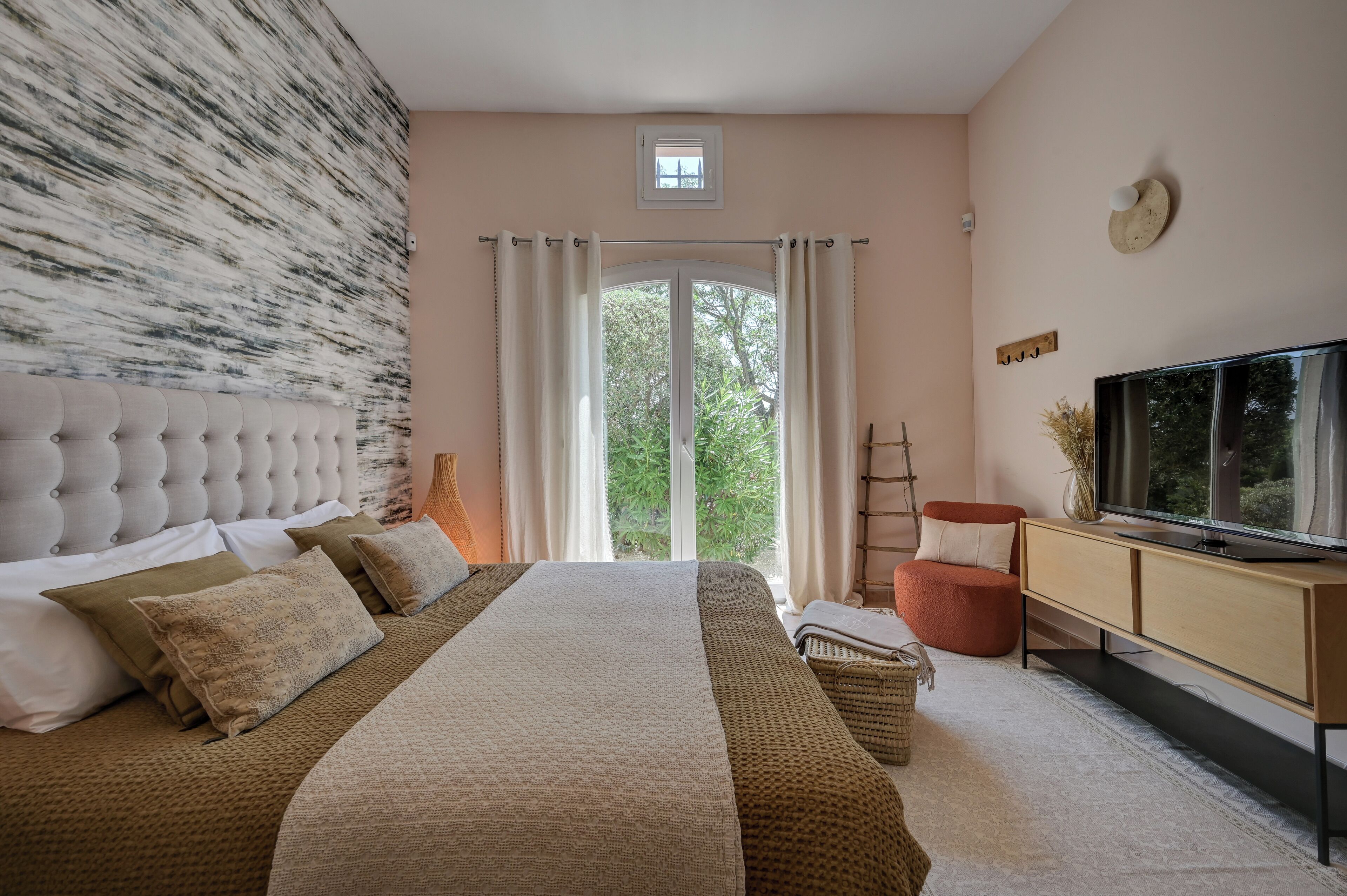 A bedroom with a large bed, a television, a window with curtains, and a view of greenery outside.