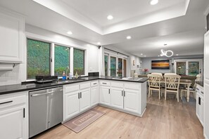 A modern kitchen with white cabinetry, a dishwasher, a dining area with a table and chairs, and a view of greenery through the windows. - Luxury Hilltop Designer House w Pool Great Views (Hacienda Heights)