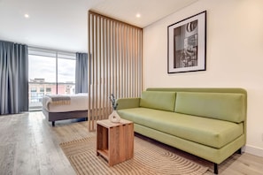 Moderni hotellihuone, jossa on vihreä sohva, puinen sivupöytä, suuri ikkuna verhoilla ja seinällä oleva kehystetty taulu.