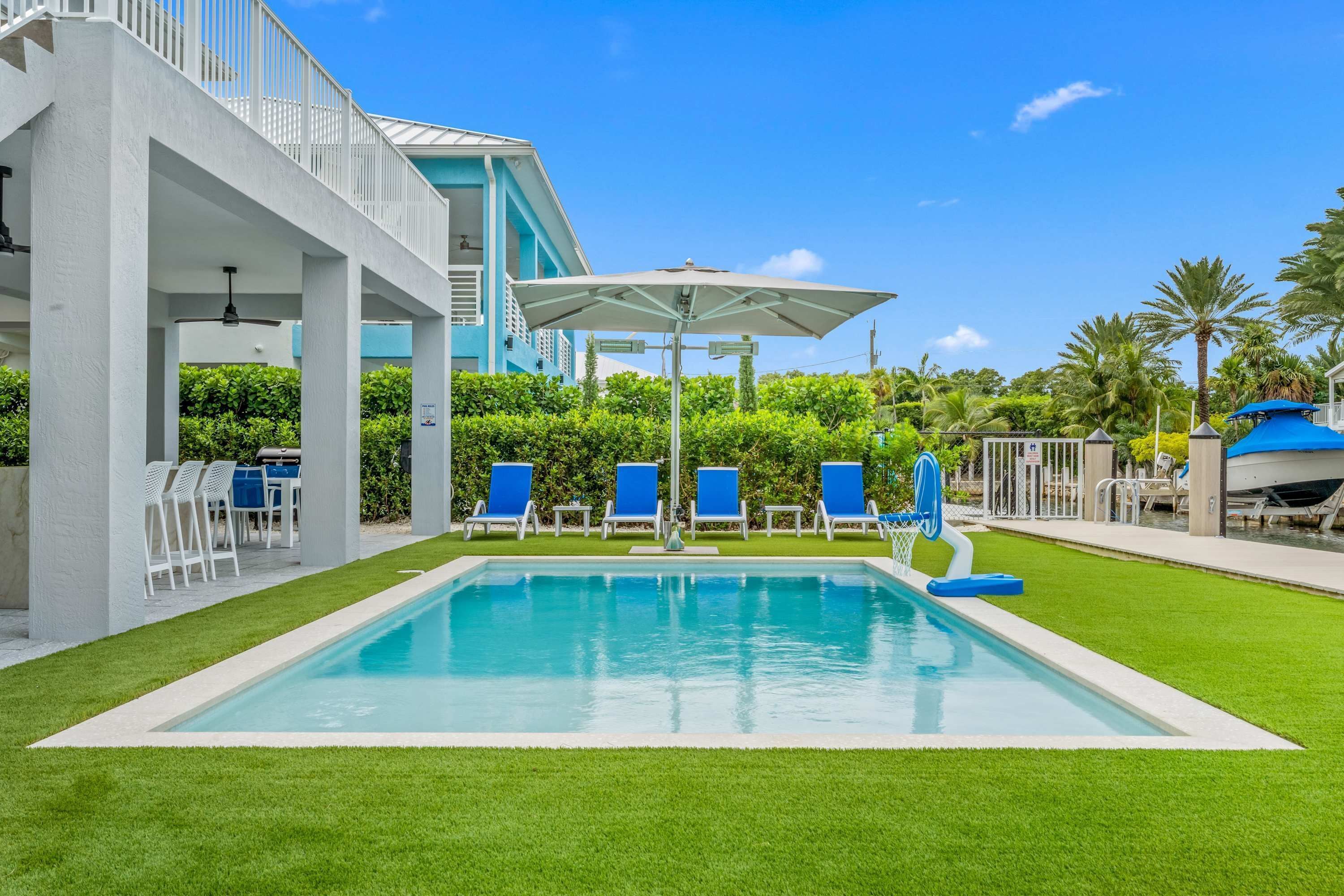 A swimming pool with blue lounge chairs and umbrellas, surrounded by well-maintained grass and palm trees.