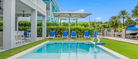 A swimming pool with blue lounge chairs and umbrellas, surrounded by well-maintained grass and palm trees.