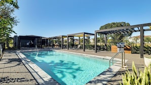 Una piscina con agua cristalina azul, rodeada de un paseo empedrado y zonas sombreadas.