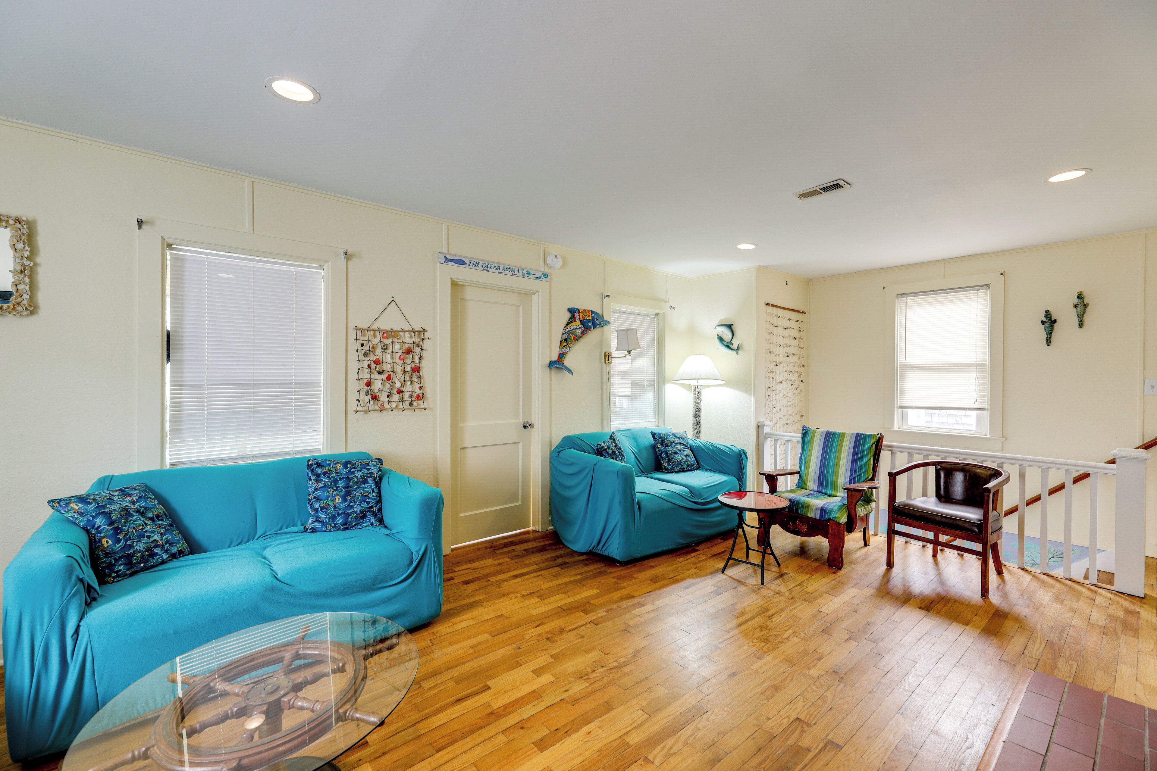 A living room with two blue sofas, a glass coffee table, wooden flooring, a staircase, and a white railing.