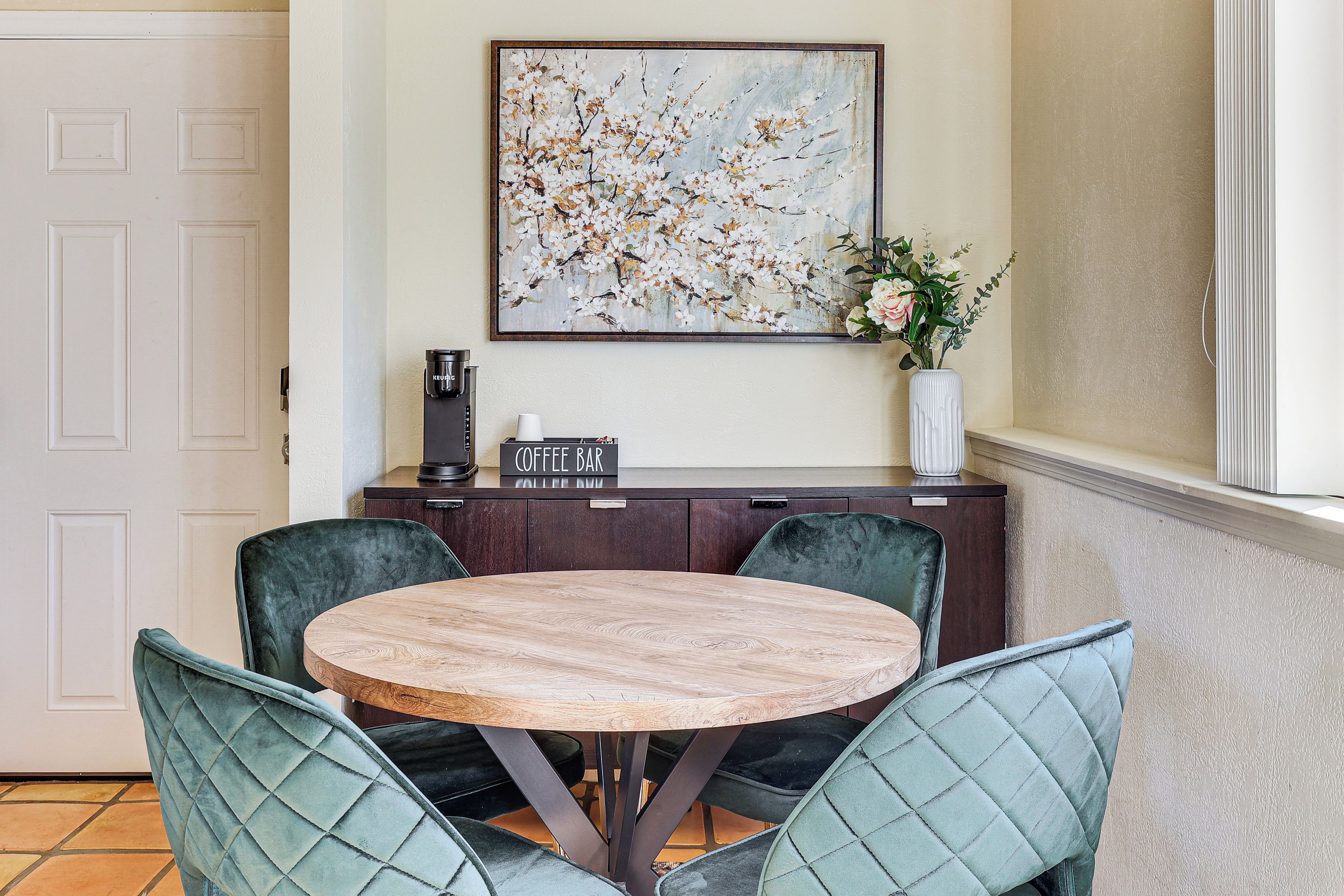 Un coin repas comprenant une table ronde en bois, quatre chaises à l’assise rembourrée de couleur verte, un bar à café et un tableau représentant des fleurs accroché au mur.