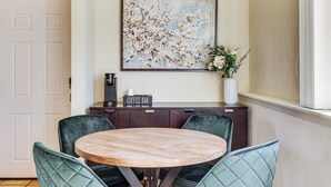 A dining area with a round wooden table, four green upholstered chairs, a coffee bar, and a floral painting on the wall.