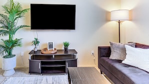 A living room with a flat-screen TV mounted on the wall, a dark-colored sofa with cushions, a floor lamp, a small side table with decorative items, and a potted plant.