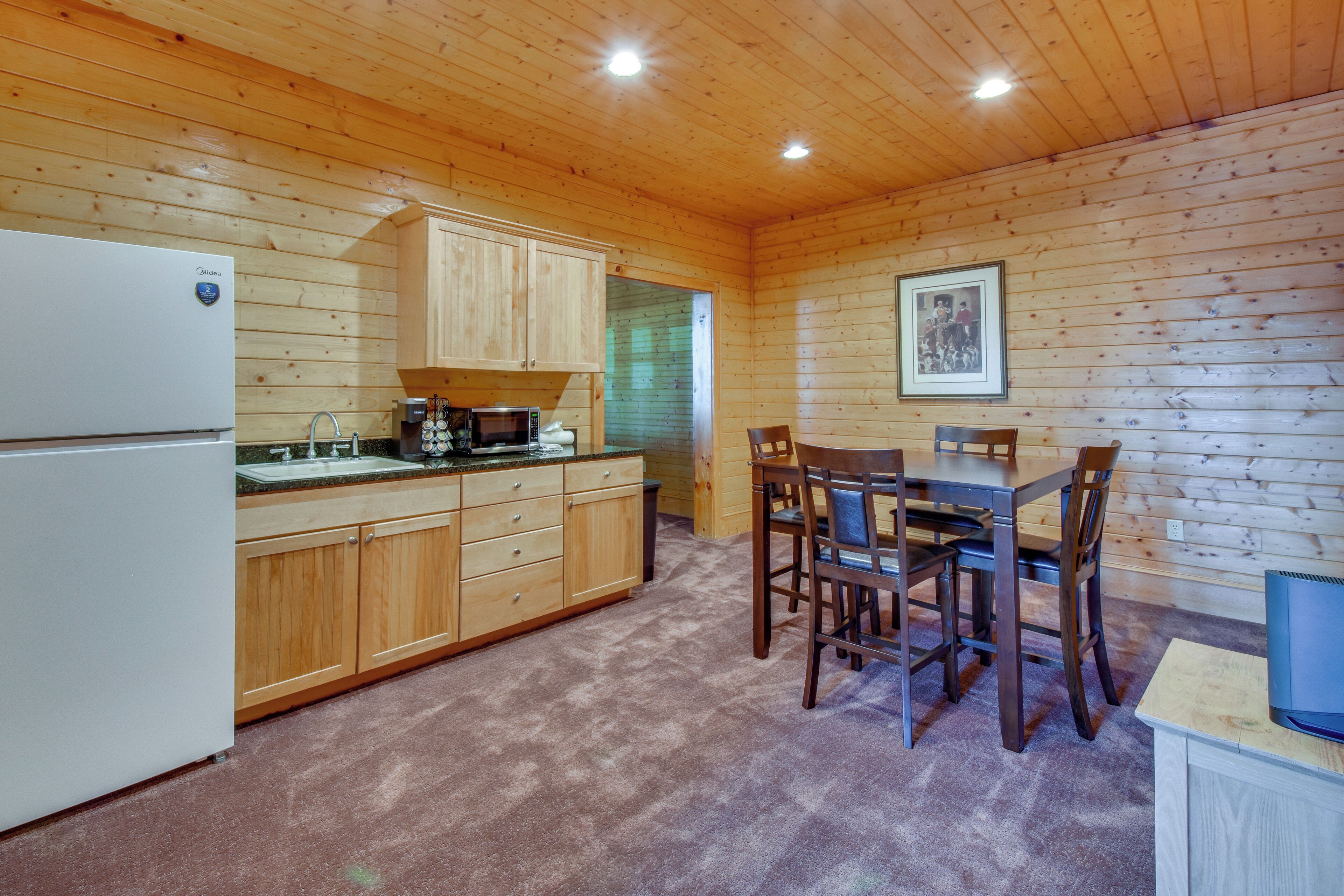 Una cucina con rivestimenti in legno, completa di frigorifero, microonde, lavandino e pensili in legno. Si nota un tavolo da pranzo con sedie e un quadro appeso alla parete.