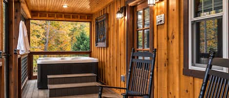 A wooden cabin with a hot tub, a wooden deck, and a view of autumn foliage.