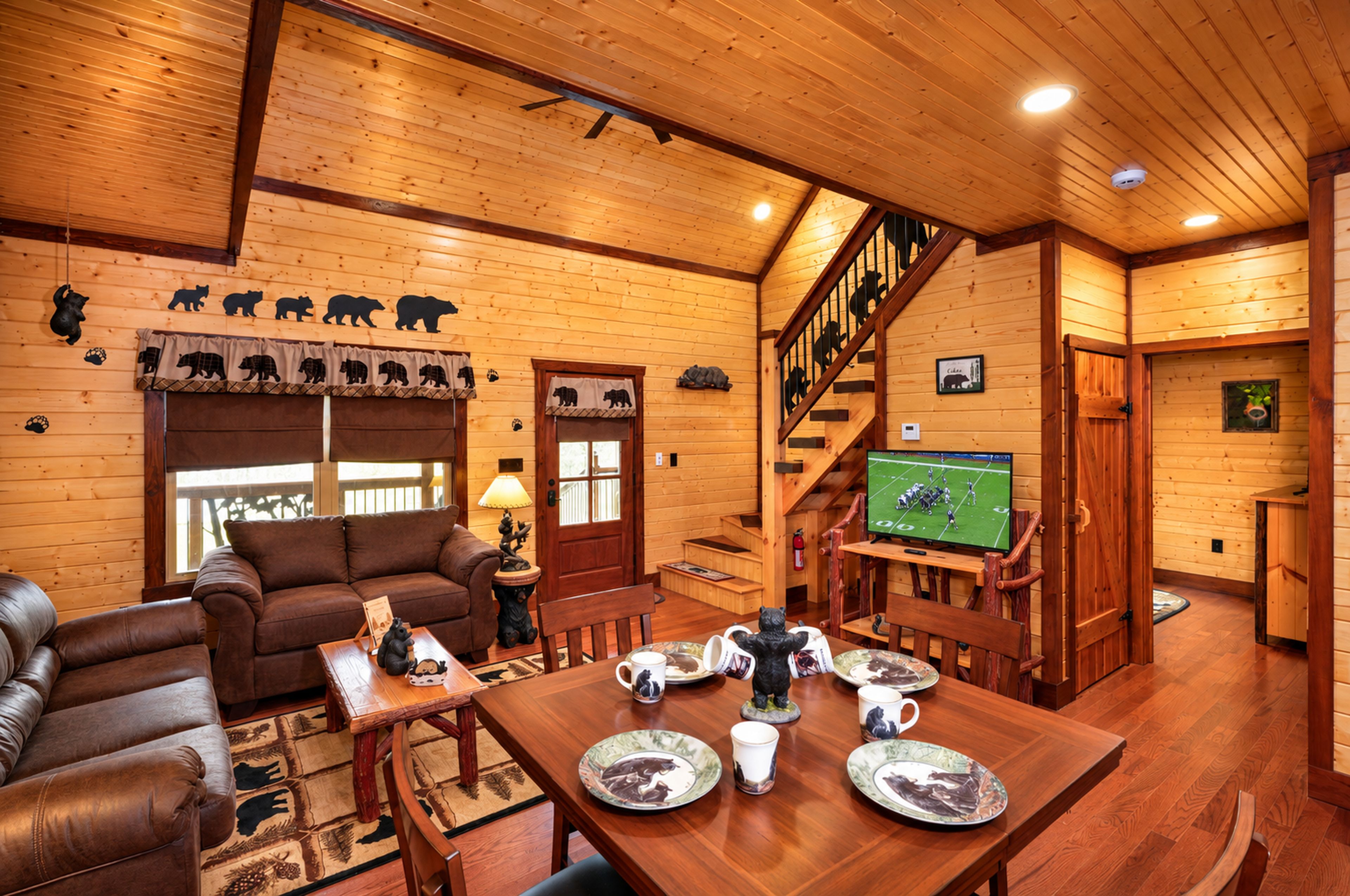 Un interior acogedor de cabaña con detalles de madera, un sofá, una mesa de comedor con vajilla temática de osos y un televisor mostrando un partido de fútbol.