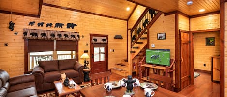 Un interior acogedor de cabaña con detalles de madera, un sofá, una mesa de comedor con vajilla temática de osos y un televisor mostrando un partido de fútbol.