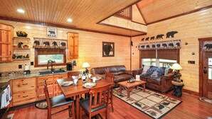 A wooden cabin interior with a dining area, a living room with a sofa and fireplace, and a kitchen with wooden cabinets and a bear-themed decor. - BEARLY LIVIN!  NEW CABIN ON 2 ACRES OF PRIVATE SMOKIES TROUT STREAM!  WOW !! (Cosby)