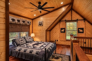 Ein Schlafzimmer einer Holzhütte mit einem Bett, einem Deckenventilator, einem Fenster und einer Bettdecke mit Bärenmotiv.
