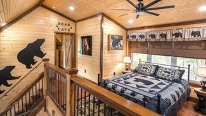 A wooden cabin interior with a bed, a ceiling fan, a sign reading "Bear Cave," and bear-themed decor. - BEARLY LIVIN!  NEW CABIN ON 2 ACRES OF PRIVATE SMOKIES TROUT STREAM!  WOW !! (Cosby)