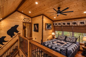 Een interieur van een blokhut met een houten plafond, een bed met een sprei met berenprint, een trap en een bordje waarop 'BEAR CAVE' staat.