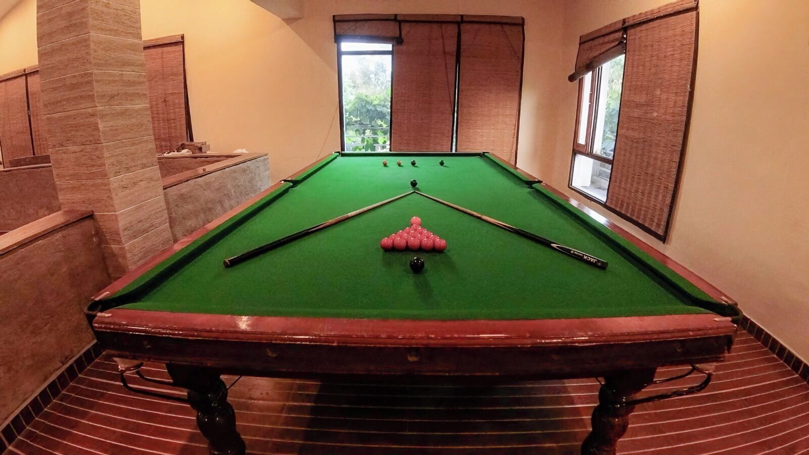 A pool table with green felt, surrounded by walls with a stone-like texture, and windows with brown curtains.