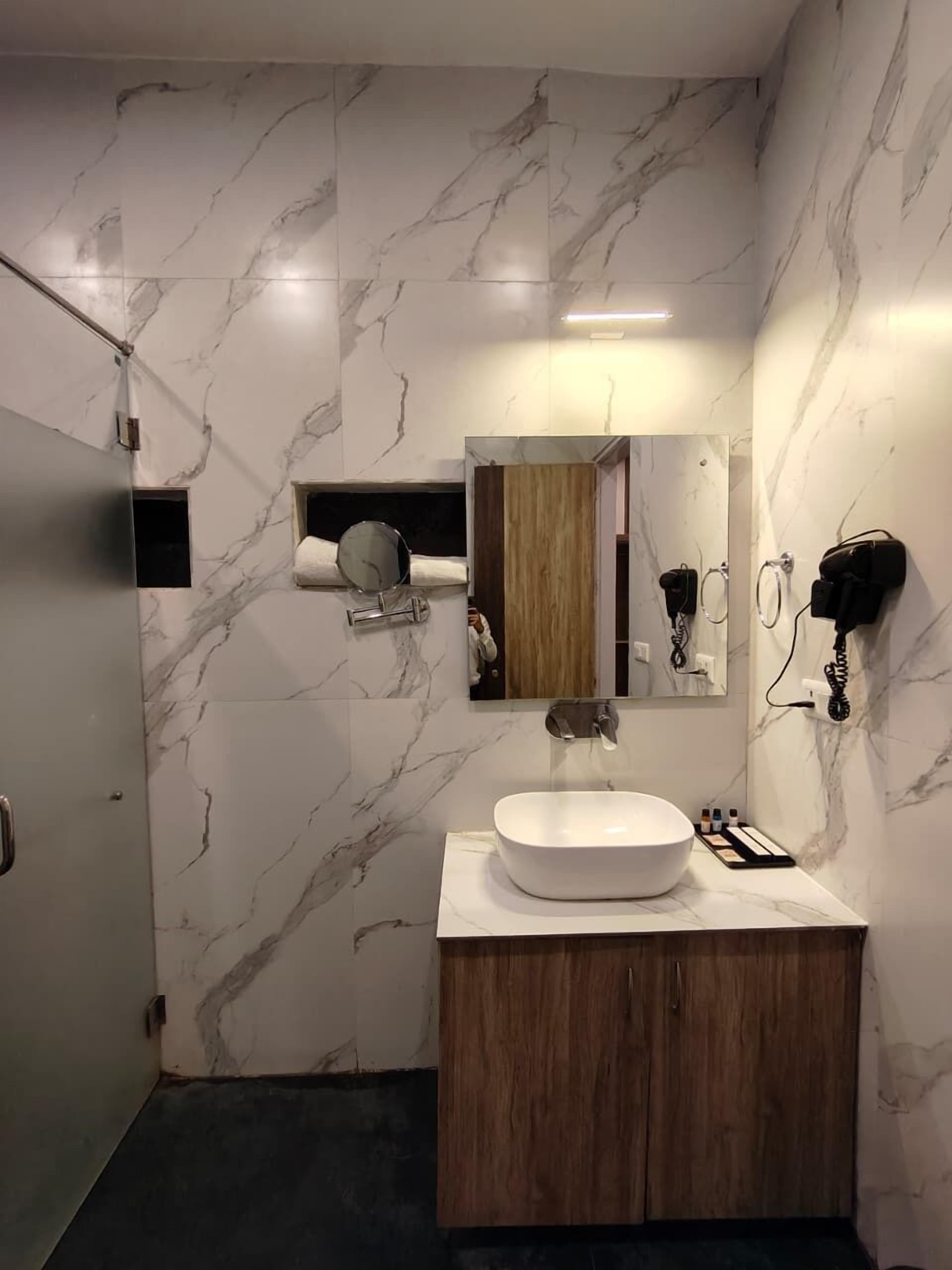 A bathroom with a marble wall, a wooden vanity, a large mirror, and a hairdryer on the wall.