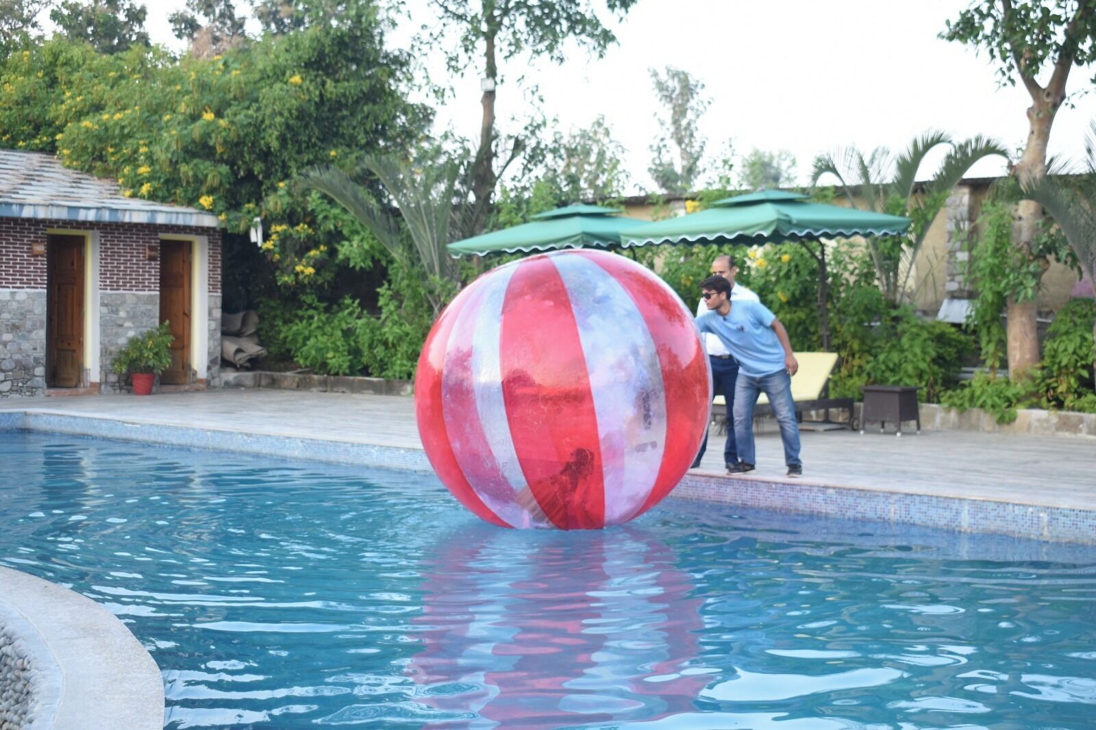 Una piscina con una gran pelota a rayas, un edificio de ladrillo y follaje al fondo.