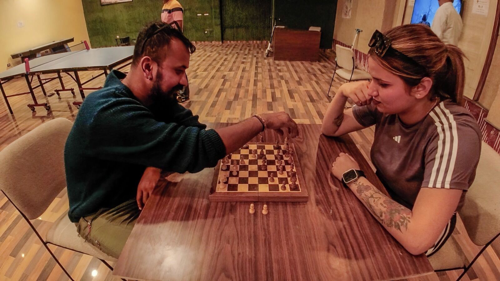Two people playing chess at a wooden table in a room with patterned flooring and a green wall.