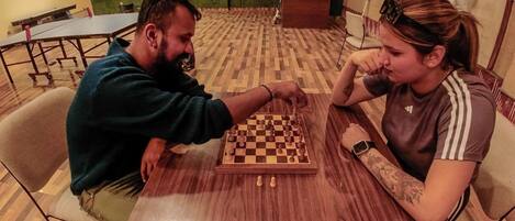 Two people playing chess at a wooden table in a room with patterned flooring and a green wall.
