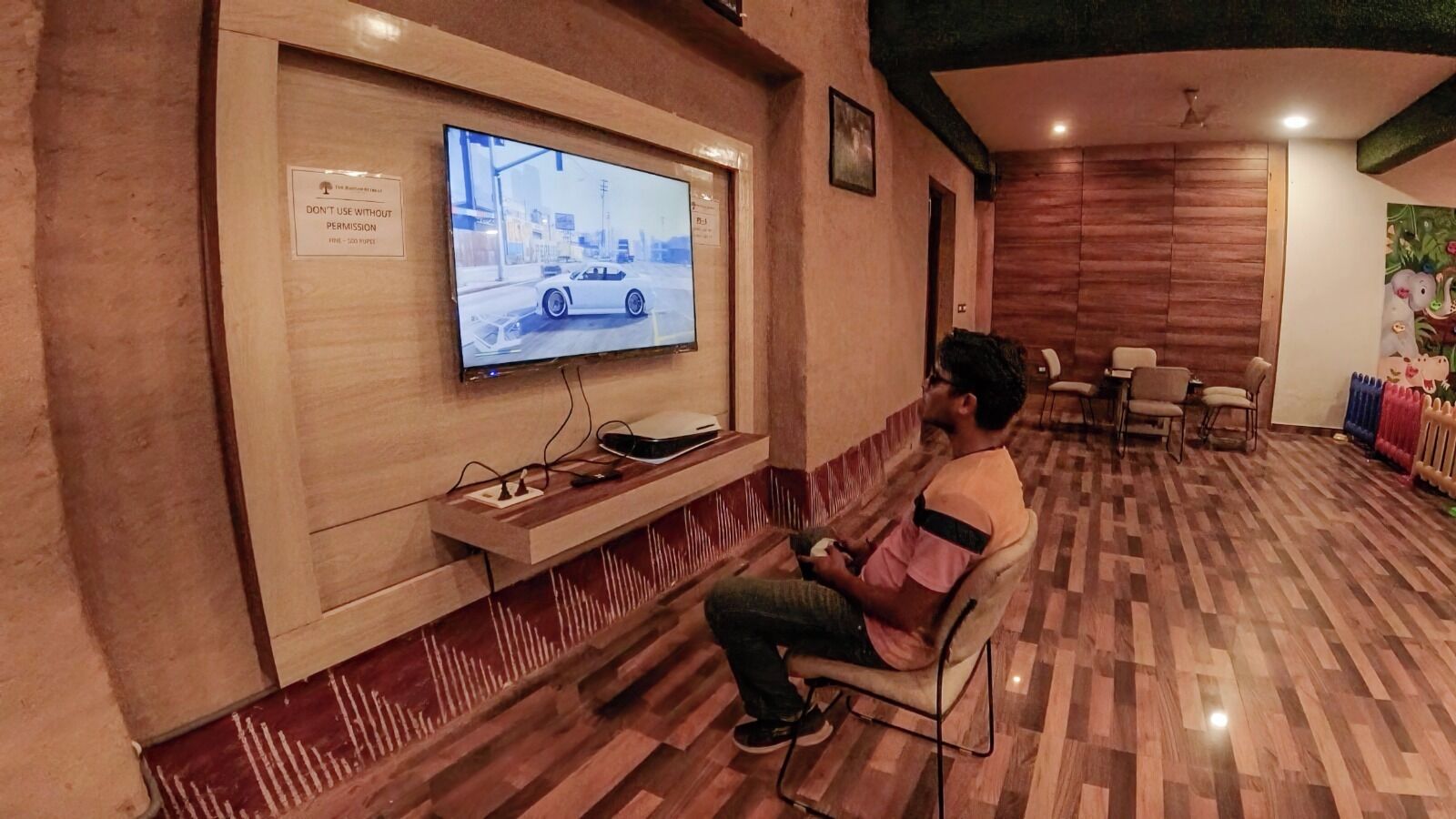A man sitting on a chair watching a TV mounted on a wall in a room with wooden flooring and a ceiling fan.