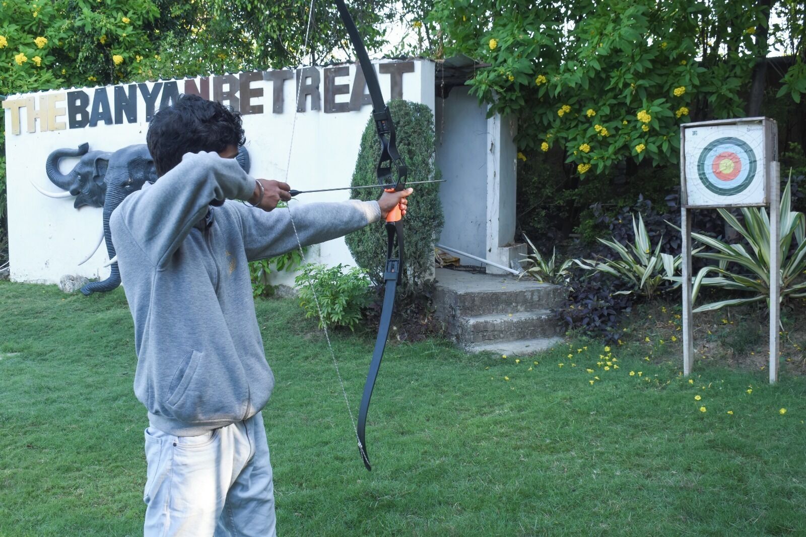 A person aiming a bow at a target in a grassy area with a sign reading 'BANYAN RETREAT' in the background.