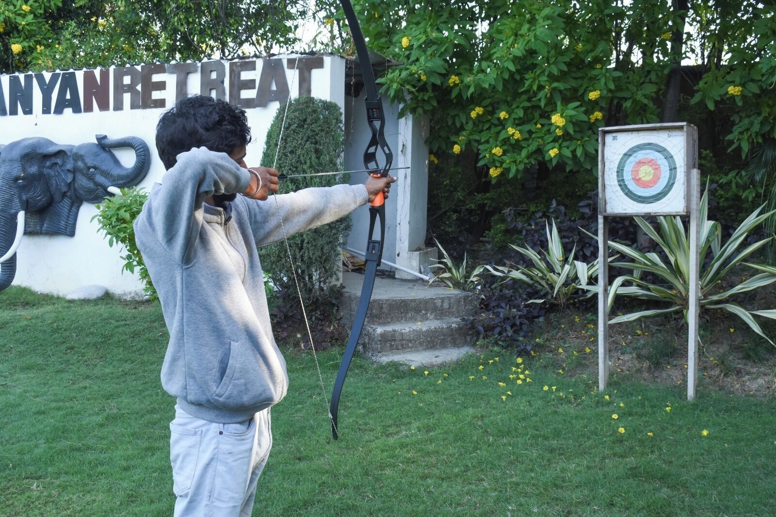 A person aiming a bow at a target in a garden with a wall that has the text 'ANYANRETREAT' and a sculpture of an elephant.