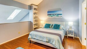 A bedroom with a bed, bedside tables, a shelf unit, and a painting of a beach scene. - Golf, Gamble & Deep-sea Fish: Little River Condo (Little River)