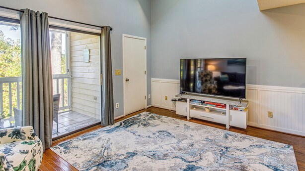 A living room with a large flat-screen TV, a white entertainment unit, a patterned rug, and a sliding glass door leading to a balcony. - Golf, Gamble & Deep-sea Fish: Little River Condo (Little River)
