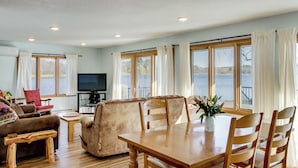 Un salón espacioso con una mesa de comedor de madera, asientos cómodos, un televisor y ventanas grandes con vistas a un cuerpo de agua.
