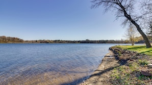 Vista despejada de un lago con orilla arenosa, un árbol sin hojas y una línea de árboles en la distancia.