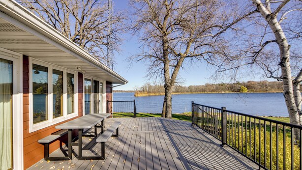 Una terraza de madera con mesas de picnic, con vista a un lago rodeado de árboles y un edificio al fondo.