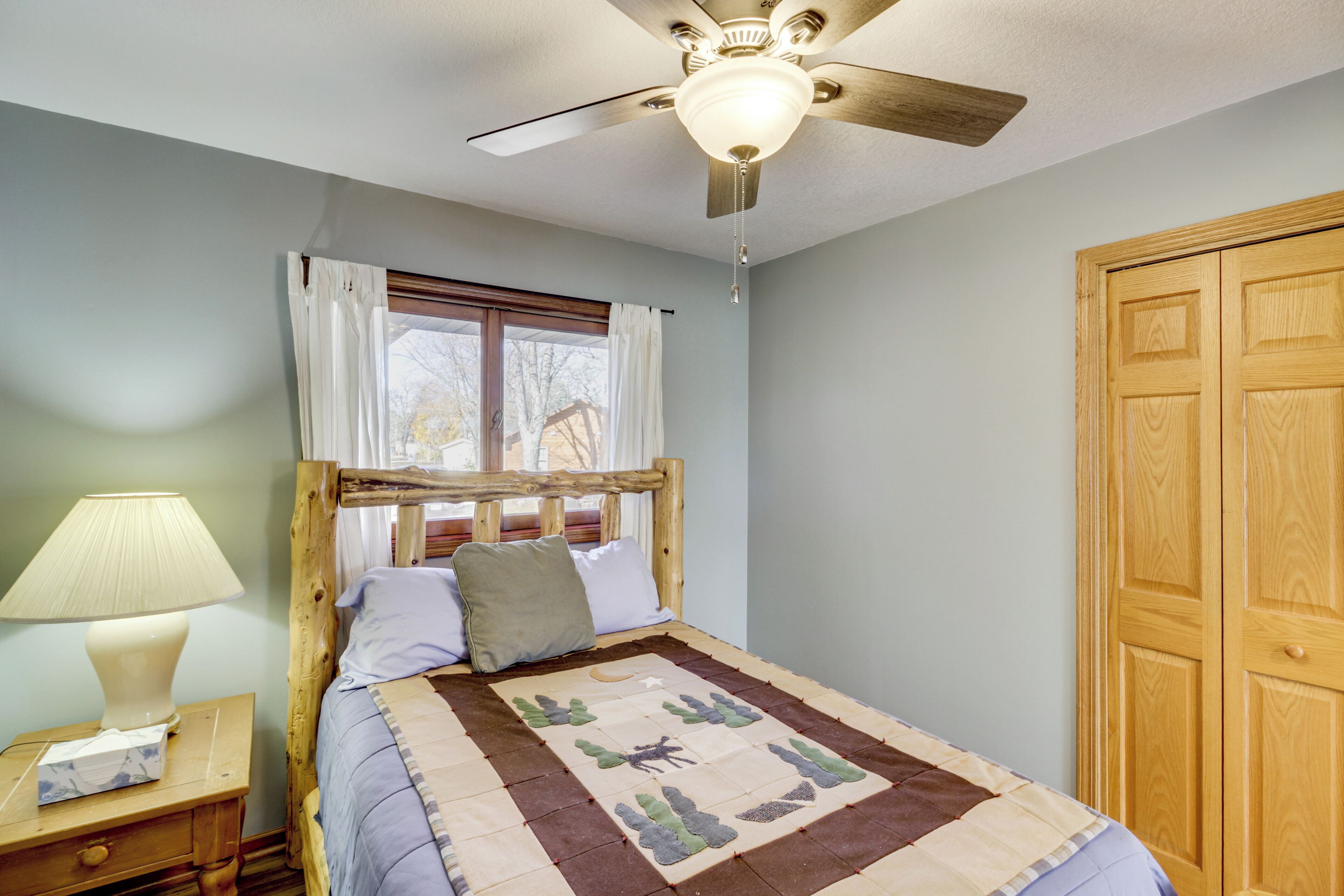 A bedroom with a wooden bed, a nightstand with a lamp, a ceiling fan, and a window with curtains.