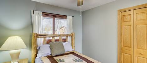 A bedroom with a wooden bed, a nightstand with a lamp, a ceiling fan, and a window with curtains.