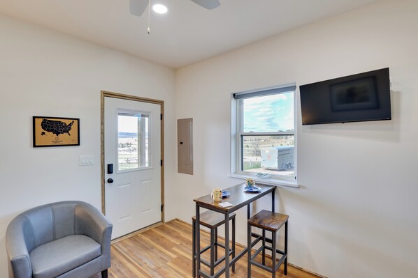 A room with a door, a window with a view, a TV mounted on the wall, and a small table with two stools.