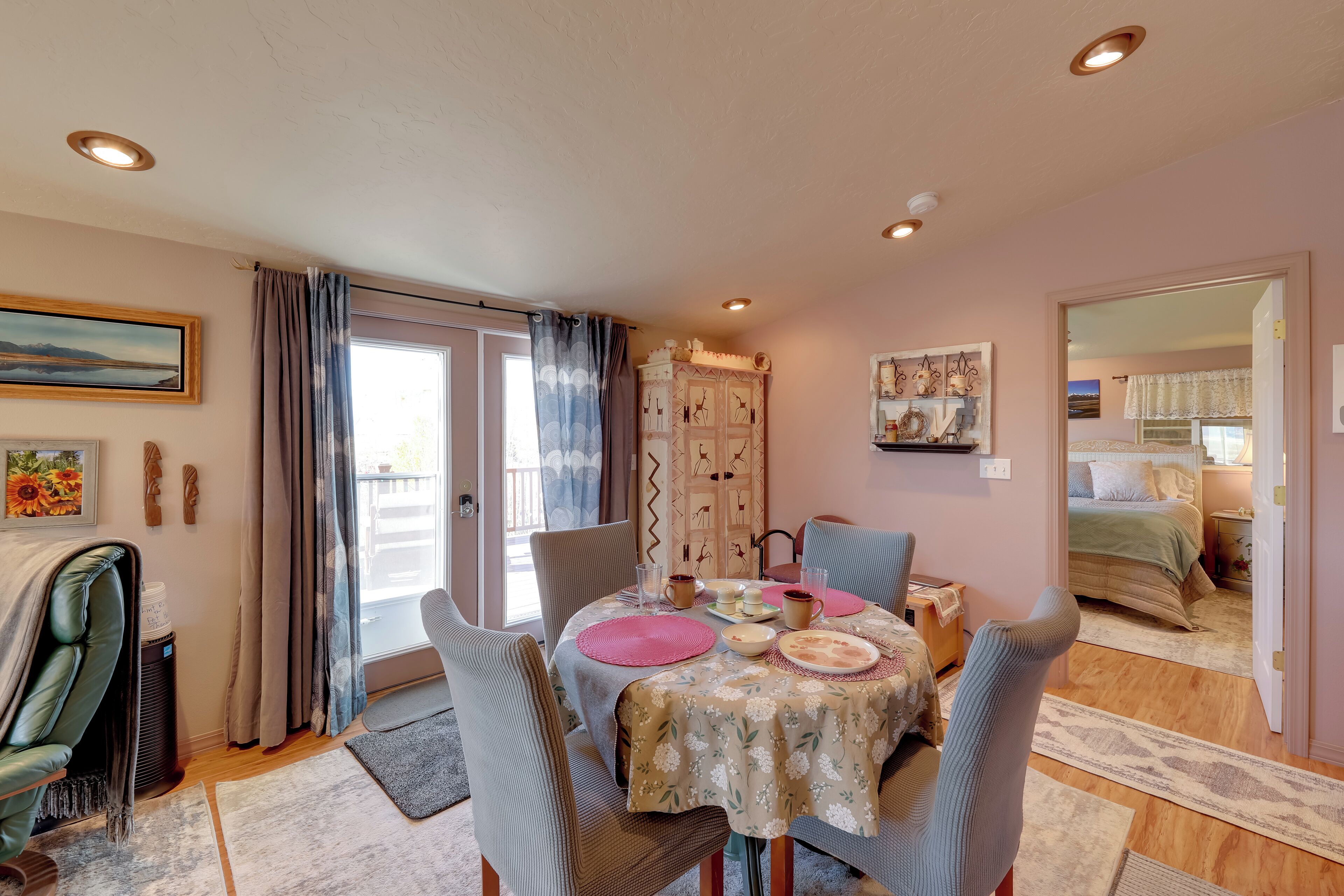 A dining area with a table set for four, chairs, a window with curtains, a radiator, and a view into a bedroom.