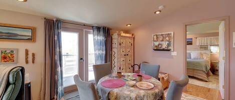 A dining area with a table set for four, chairs, a window with curtains, a radiator, and a view into a bedroom.