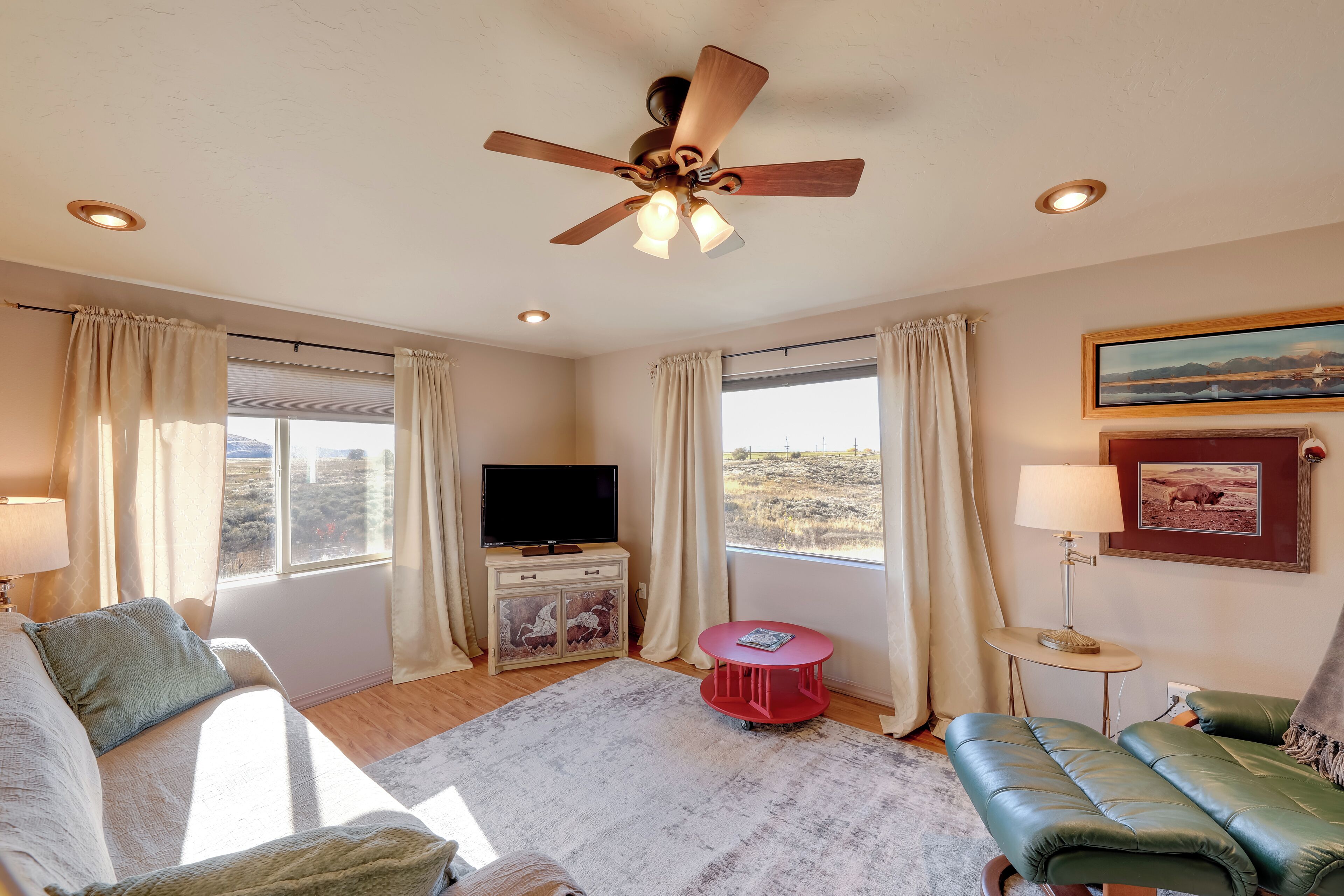 Una habitación con ventilador de techo, televisor, una mesa roja, sofá y vistas al exterior.