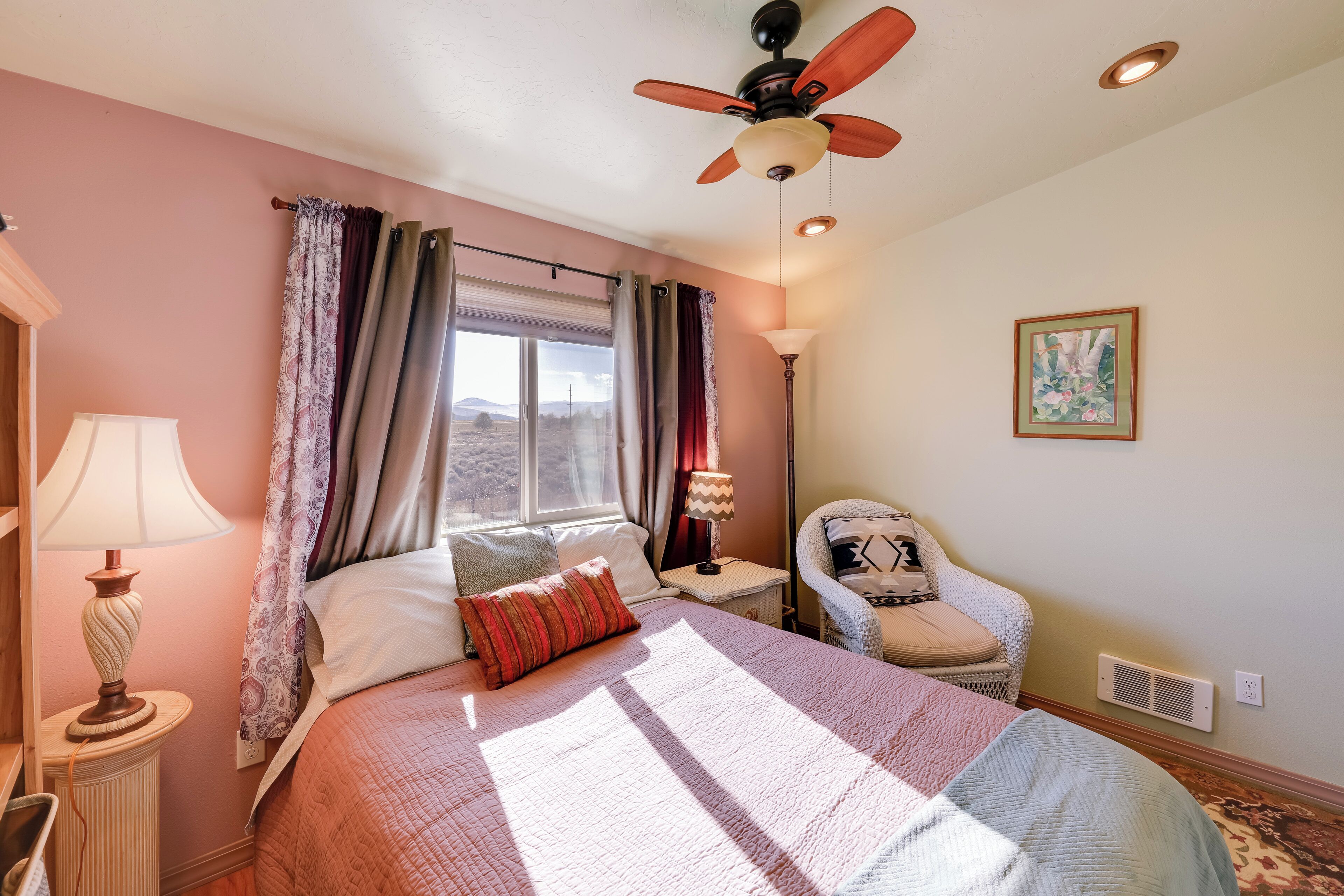Une chambre à coucher avec un lit, une chaise, une fenêtre avec des rideaux, un ventilateur de plafond et une lampe.