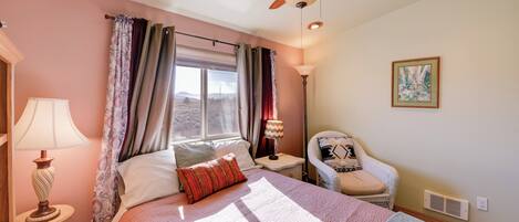 Una camera da letto con un letto, una sedia, una finestra con tende, una ventola a soffitto e una lampada.