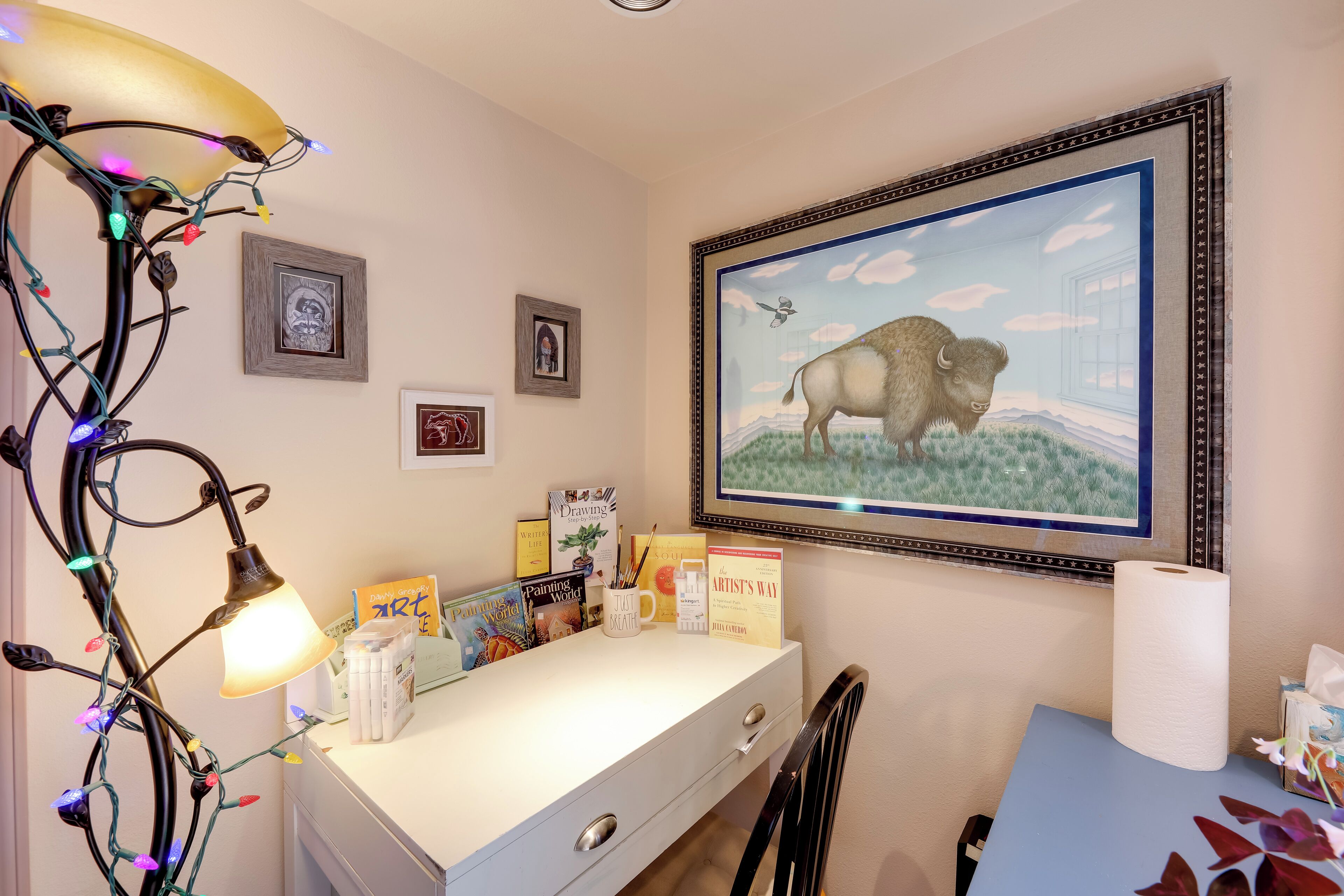 A room with a desk, a chair, a framed painting of a bison, and a lamp with colorful lights.