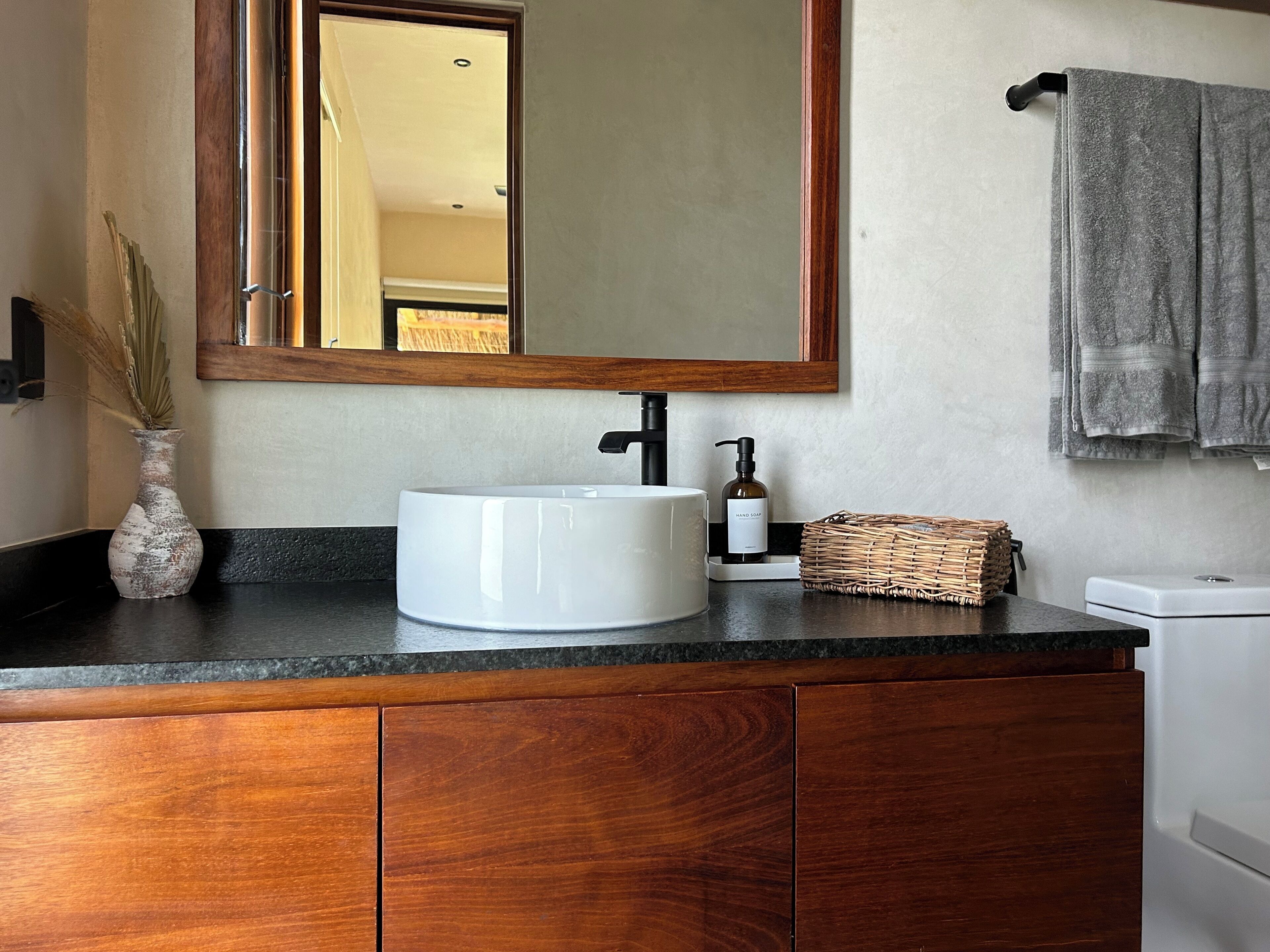 Une salle de bain avec un lavabo blanc, un meuble-lavabo en bois, un miroir, un flacon de savon liquide et un panier en osier.