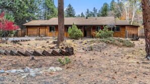 A two-story log cabin with a front porch, surrounded by pine trees and a dry, rocky landscape.