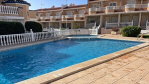 Una piscina con solárium empedrado, rodeada por una valla blanca y un edificio con balcones.