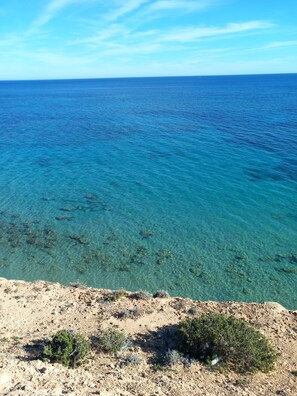 Paisagem costeira com mar azul cristalino, terreno rochoso e vegetação rala.