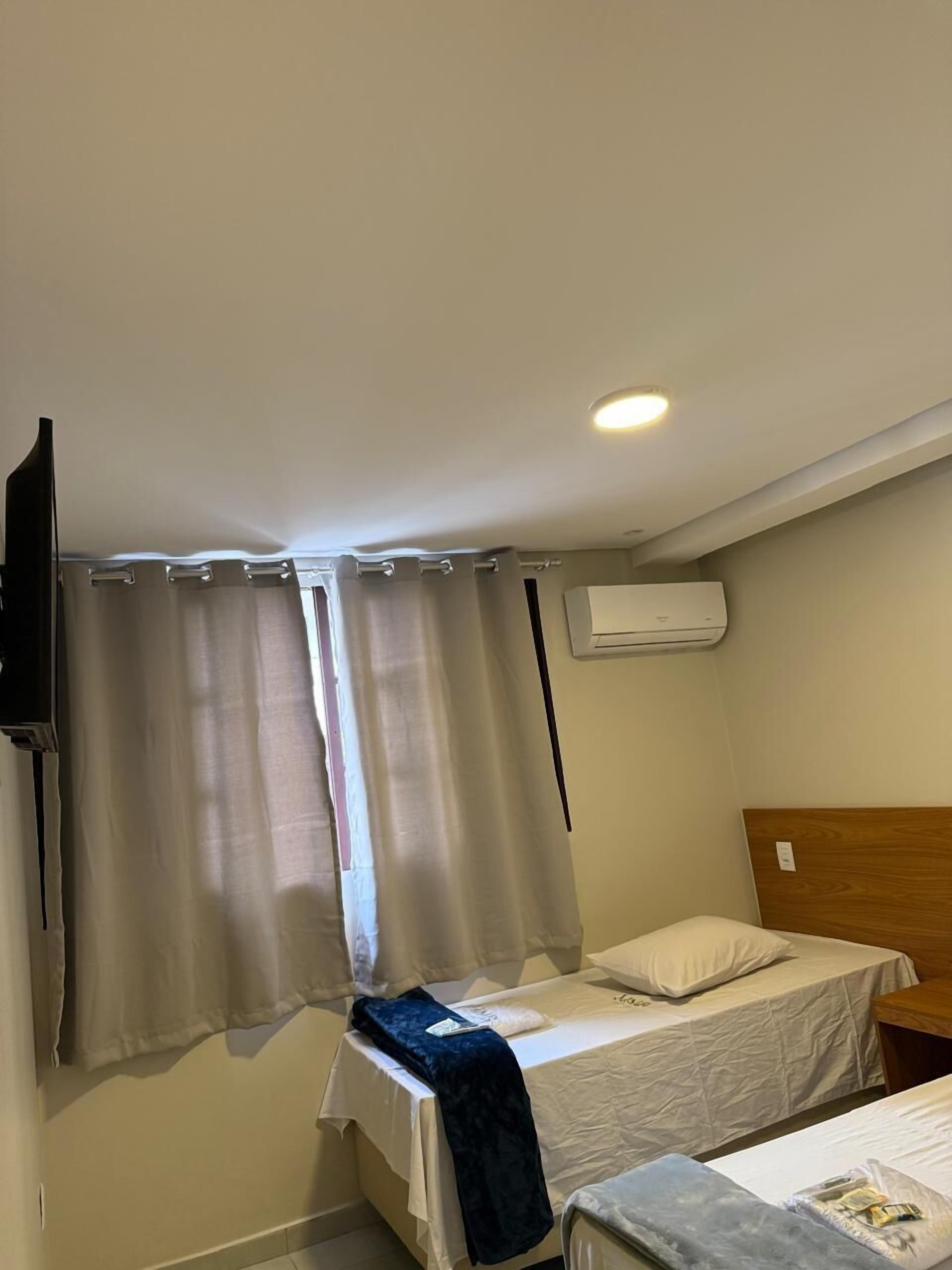 Une petite chambre avec un lit simple, une tête de lit en bois, un téléviseur à écran plat fixé au mur et une fenêtre avec des rideaux.