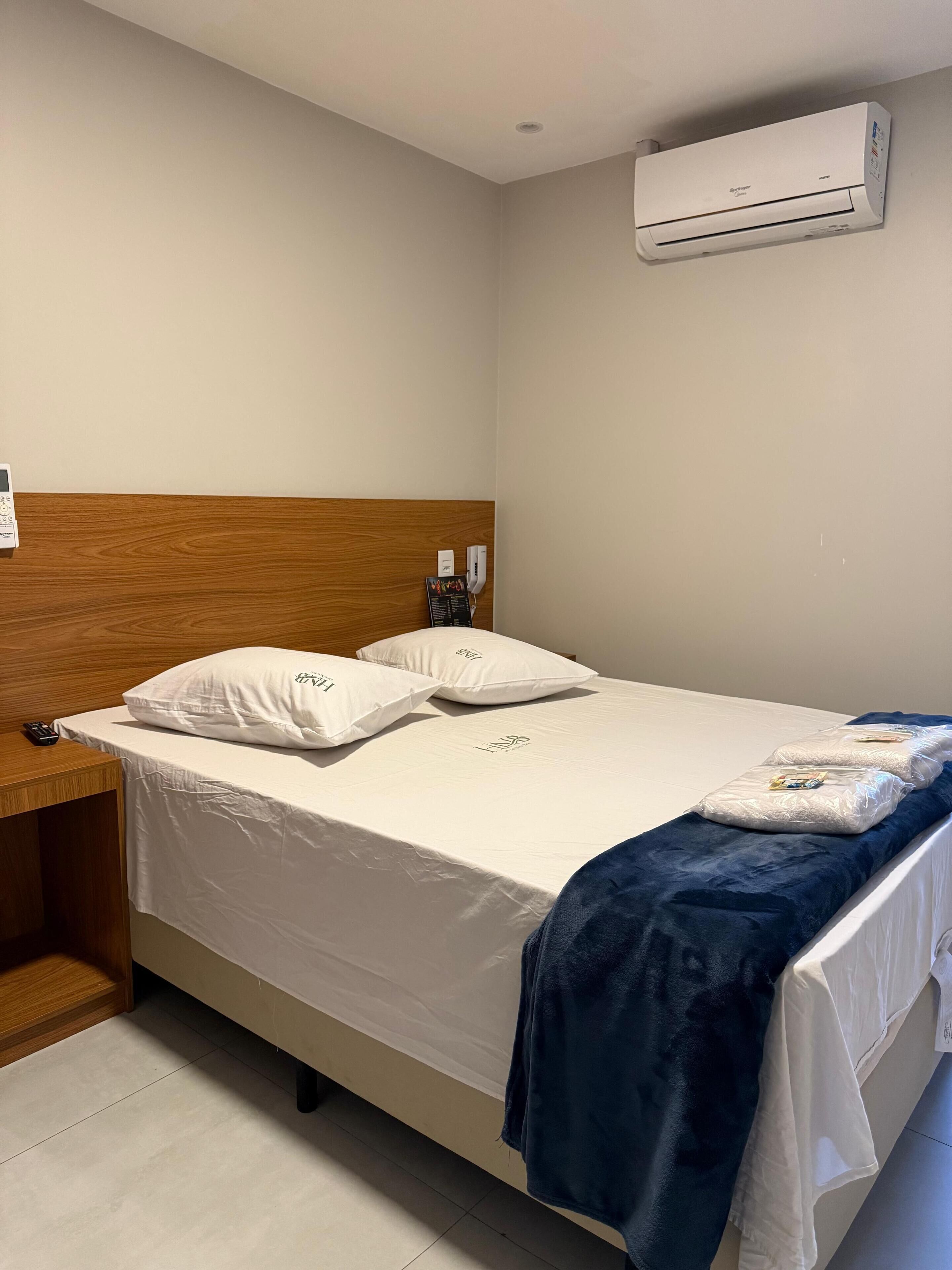 A hotel room with a bed, a wooden headboard, a bedside table with a remote control, a blue blanket, and an air conditioning unit on the wall.