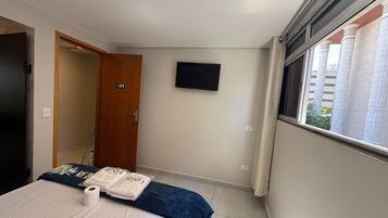A hotel room with a bed, a TV mounted on the wall, a door with a number, and a window with a view of buildings.