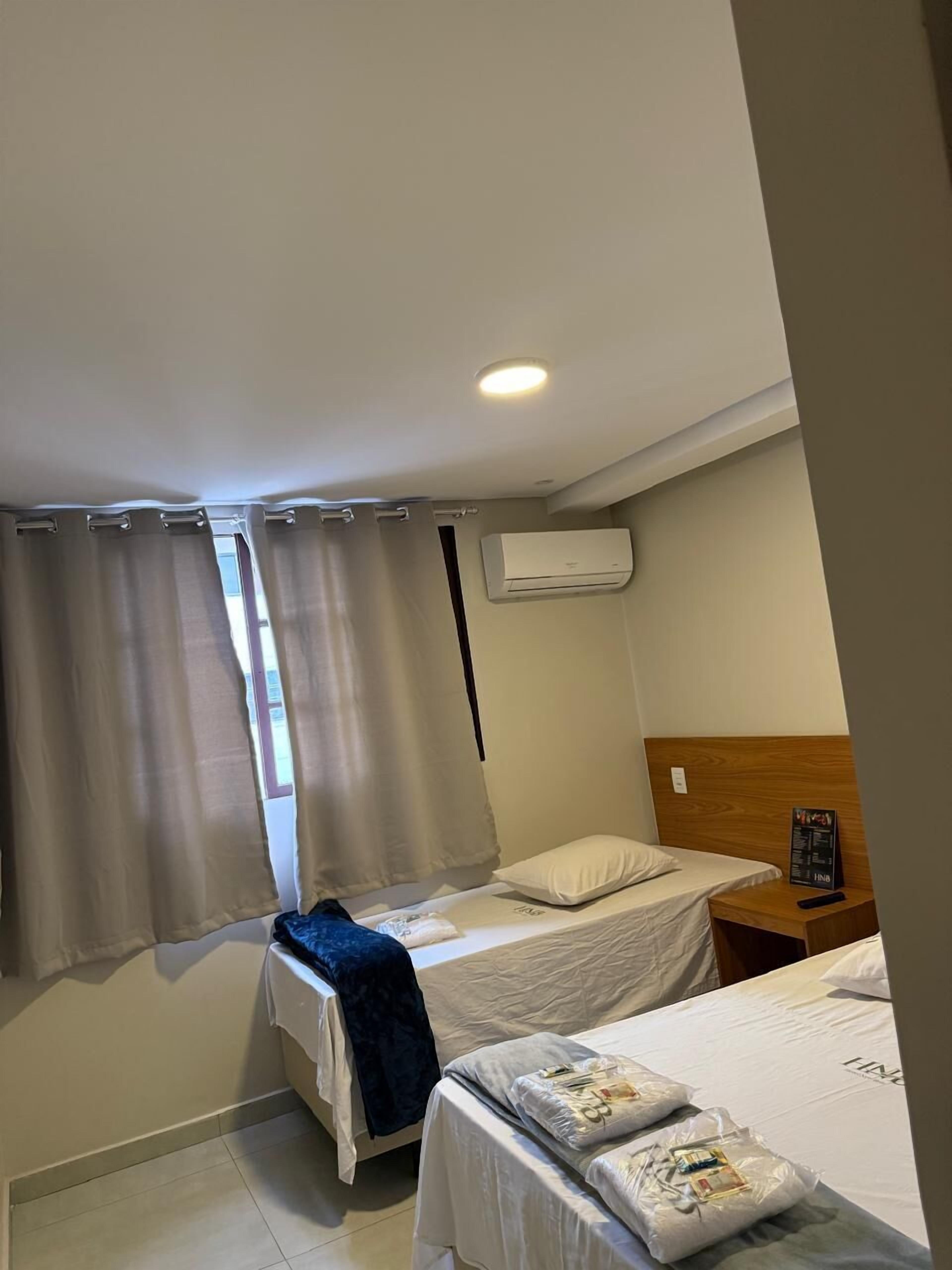 Ett hotellrum med två sängar, en sänggavel i trä, en luftkonditioneringsanläggning, ett nattduksbord med en meny och ett fönster med gardiner.
