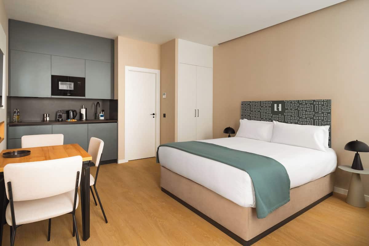 A modern hotel room with a bed, a desk, and a kitchenette.